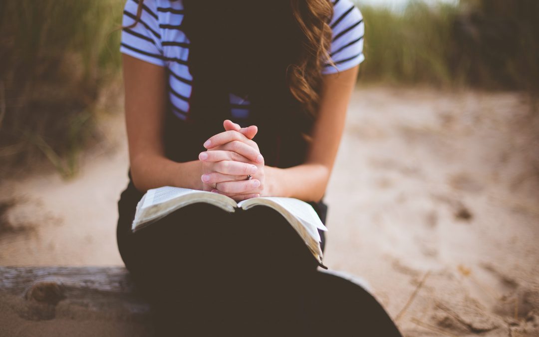 Woman sitting at the beach on a log, with tall grass in the background. Her hands are folded over her Bible and her head is bowed in prayer.