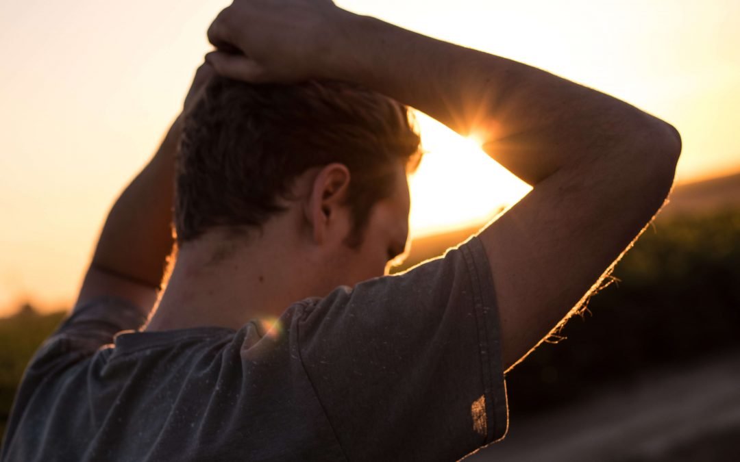A young man is watching the sunset, his hands over his head in distress.