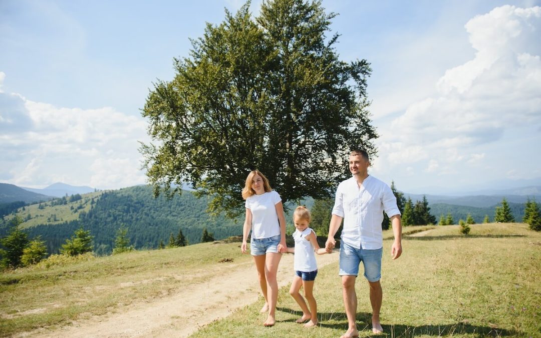 Mother, child, and father are walking barefoot on the grass with a mountain range behind them. The child is in between the parents, and the parents are each holding one of her hands.